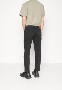 Jeans in denim nero con vestibilità slim, dotati di tasche posteriori e di un'etichetta rossa in vita. Abbinati a una t-shirt a collo rotondo beige e a sneakers nere.