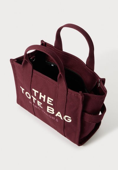 Sac fourre-tout en toile bourgogne avec double poignée, fermeture éclair et texte blanc audacieux indiquant "LE SAC FOURRE-TOUT" et "MARC JACOBS." Compartiments intérieurs visibles.