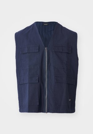 Mornarsko modra brezrokavnika iz bombaža z zadrgo spredaj, ki ima štiri sprednje žepe in obliko V-izreza. Gladka tekstura in minimalni detajli.