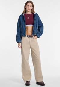 Spijkerjack, bordeaux crop top, beige wijde broek en bruine laarzen. Opvallende details zijn een trekkoord capuchon en een gestructureerde stof.