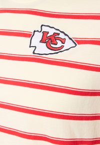 Ριγέ μπλουζάκι σε κρεμ χρώμα με κόκκινες ρίγες. Διαθέτει κεντημένο οβάλ λογότυπο με τα γράμματα "KC" σε κόκκινο πάνω σε λευκό φόντο. Μαλακή βαμβακερή υφή.