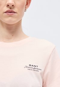 Κοντινό πλάνο ενός ατόμου που φοράει ανοιχτό ροζ μπλουζάκι με το κείμενο "GANT American Sportswear New Haven" τυπωμένο στο στήθος.