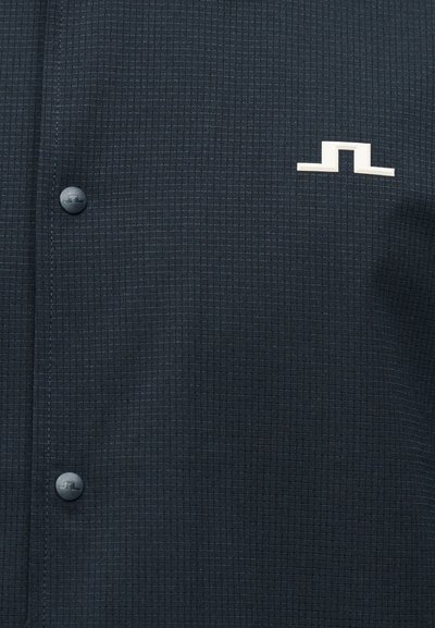 Σκούρο υφασμάτινο σχέδιο με δύο ματ κουμπιά που φέρουν ένα μικρό ανάγλυφο λογότυπο και ένα λευκό, γεωμετρικό έμβλημα ραμμένο κοντά στο πάνω δεξιά μέρος.