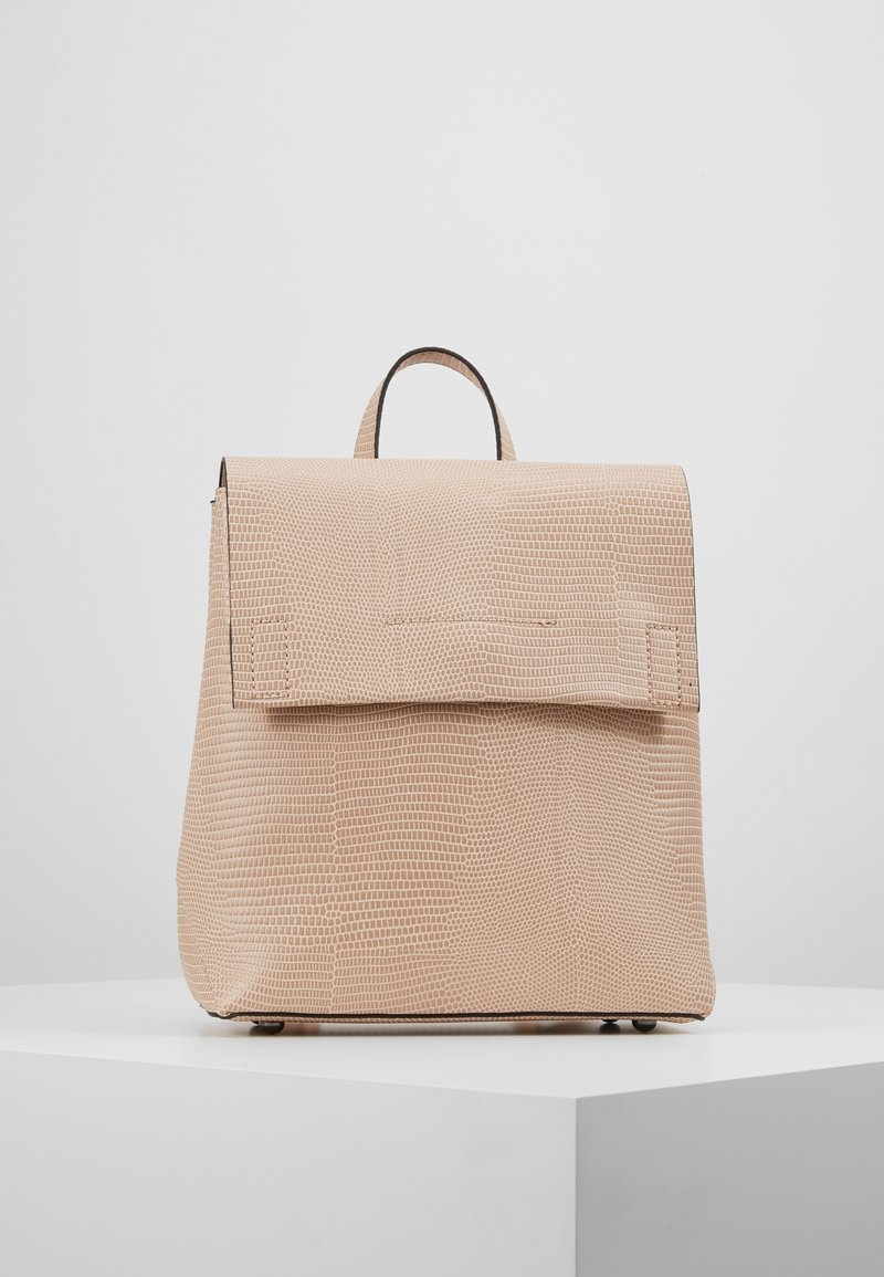 Sac à dos beige texturé avec poignée supérieure et grande fermeture à rabat, posé sur une plateforme blanche contre un fond gris clair uni.