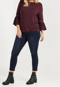 Viininpunainen paita liehuvine hihoineen ja pitsikoristeineen, yhdistetty tummansinisiin kapealahkeisiin farkkuihin ja mustiin nilkkureihin, joissa on paksu korko.