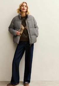 Gevoerde geblokte jas in bruin en crème, met een hoge kraag en zakken aan de voorkant, gecombineerd met een donkerbruine trui en wijde denimbroek.