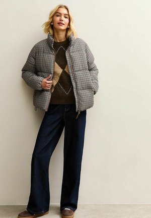 Giacca imbottita a quadri marrone e crema, con colletto alto e tasche frontali, abbinata a un maglione marrone scuro e pantaloni in denim a gamba larga.