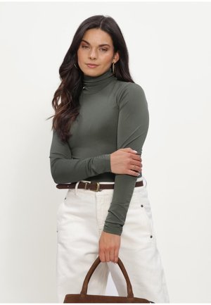 Woman with long dark hair wears olive green turtleneck and white pants, holding brown handbag with both hands in front of her.