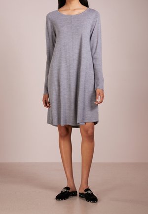 Femme portant une robe grise ample à manches longues et des chaussures noires à enfiler avec des ornements décoratifs, debout sur un sol uni.