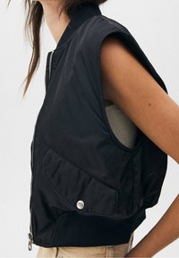 Schwarze ärmellose Jacke mit Reißverschluss vorne, geripptem Kragen und großer Seitentasche mit Druckknopfverschluss. Glatte Textur, leichtes Material.