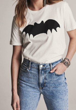 Kobieta w białym t-shircie z dużą czarną grafiką nietoperza, niebieskich jeansach z wysokim stanem, srebrnych bransoletkach z łańcuszka i długich kolczykach.