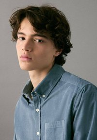 Skräddarskjord ljusblå kordflätskjorta med knapp-down krage, utrustad med en bröstficka och subtil textur, bärs av en person med vågigt hår.