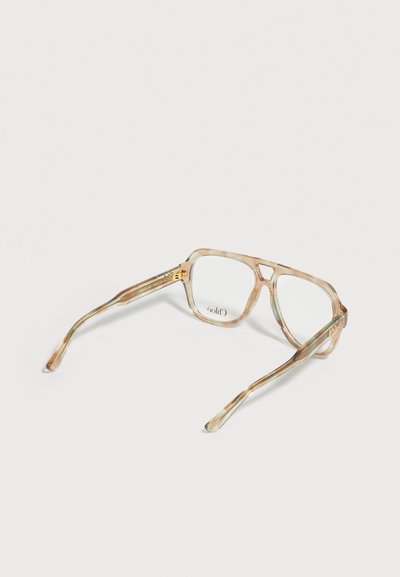 Transparente beige und grün gemusterte Schildpatt-Brille mit quadratischen Gläsern, die mit der Vorderseite nach unten auf einer weißen Oberfläche liegt.