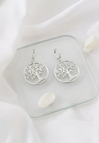 Orecchini a forma di albero in argento con un intricato design a intaglio, caratterizzati da dettagli a foglia, sospesi da ganci a leva su un'esposizione in vetro.