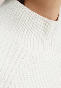 Bílý pletený rolák s žebrovanou texturou a vysokým límcem, záběr zblízka zvýrazňující vzor stehů.