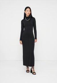 Czarna, dopasowana sukienka midi z długim rękawem, wysokim kołnierzem i detalami szwu z przodu; noszona z czarnymi sandałami na cienkich obcasach.