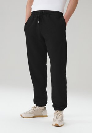 Személy fekete, elasztikus derékpántos, zsebes melegítőnadrágot és bézs színű sportcipőt visel, egyszerű világos háttér előtt áll.