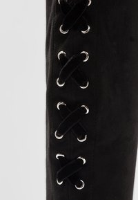 Botte haute jusqu'au genou en daim noir avec un laçage latéral orné d'œillets en métal argenté et de lacets noirs croisés pour un ajustement réglable.