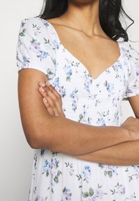 Kvinna med armarna i kors, iklädd en vit klänning med blå och lila blommönster och puffärmar.