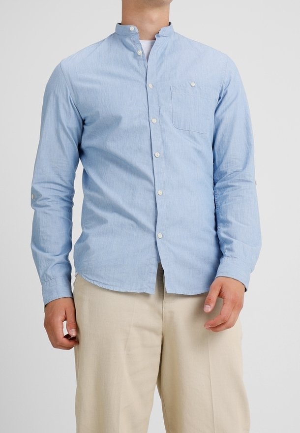 Homme portant une chemise bleu clair à boutons avec col mandarin, un sous-pull blanc et un pantalon beige sur fond uni.
