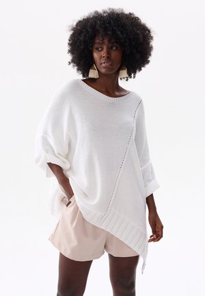Vrouw met krullend haar, die grote kwastoorbellen draagt, een losse witte asymmetrische gebreide trui en beige korte broek, staand met één hand in haar zak.