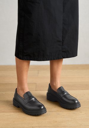 Personne portant des mocassins chunky noirs et une jupe midi noire debout sur un sol en bois, jambes visibles à partir du milieu du mollet vers le bas.