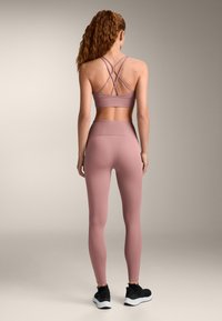 Femeie cu păr creț roșcat, purtând un sutien sportiv mov cu spatele în cruce, colanți cu talie înaltă și pantofi sport negri, stând cu fața spre spate.