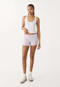 Helle lila Sportshorts mit einem breiten Bund, kombiniert mit einem weißen Tanktop. Das Modell trägt silberne Sportschuhe mit weißen Socken.
