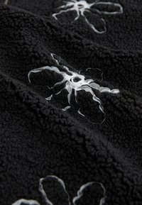 Sort fleece-stof med hvide broderede blomsterdesigns. Teksturen er blød og dunet med hævede fibre for ekstra dybde.