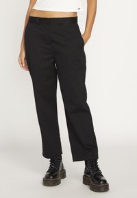 Pantalon en coton noir à coupe droite, avec poches latérales et devant plat. Associé à des bottes noires à lacets pour un contraste.