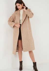 Manteau croisé en tissu beige texturé, avec de larges revers, des manches longues et des poches latérales. Stylisé sur un pull léger et une jupe.
