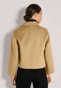 Frau mit dunklem Haar im Dutt, trägt einen kurzen kamelfarbenen Mantel und schwarze Hosen, mit dem Rücken zu einer einfarbigen hellen Kulisse.