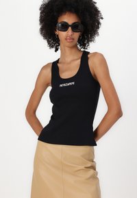 Melnas ribotā auduma bezpiedurkņu krekls ar balto tekstu "PATRIZIA PEPE". Modelis valkā beige ādas svārkus un melnas taisnstūra formas saulesbrilles. Vienkāršs dizains.