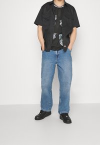 Camisa negra de manga corta sobre una camiseta de malla gráfica negra, combinada con jeans de mezclilla sueltos de color azul claro y zapatillas negras. Tejidos texturizados y ajuste casual.