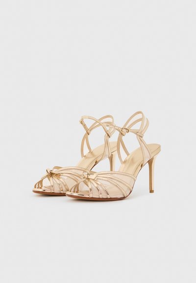 Χρυσά ψηλοτάκουνα σανδάλια με λουράκια και αγκράφα στον αστράγαλο και ανοιχτές μύτες, τοποθετημένα πάνω σε απλό ανοιχτό γκρι φόντο.