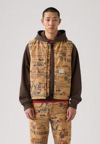 Smeđi prsluk s otisnutim grafičkim motivima, koji prikazuje prizore i tekst, preko tamnog dukserice. Nosiv s odgovarajućim hlačama s tiskom, stvarajući usklađen outfit.