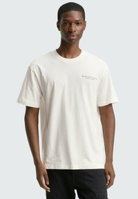 Crème katoenen t-shirt met korte mouwen, ronde hals en grafische tekst op het bovenste linker borstgebied. Model draagt donkere broek.