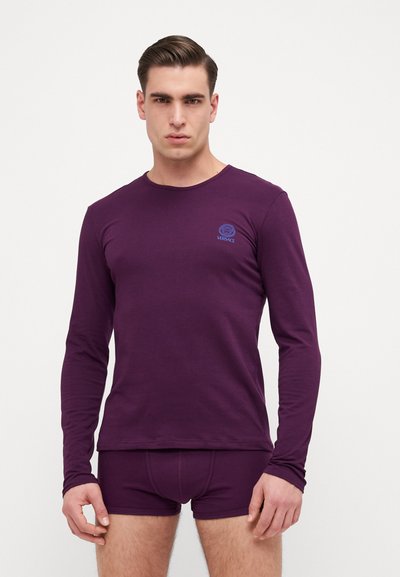 Lila Langarmshirt mit Rundhals, mit einem blauen Logo auf der linken Brust; kombiniert mit passenden lila Unterhosen. Glatte Textur.