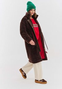 Žena v zelené čepici, dlouhém hnědém fleece kabátu, červené mikině, bílých kalhotách a hnědých teniskách, jde s mírným úsměvem.