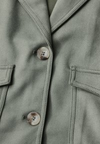 Grüne Stoffjacke mit strukturiertem Finish, einem Knopfverschluss und zwei aufgesetzten Taschen auf der Vorderseite. Die Knöpfe sind hell und rund.