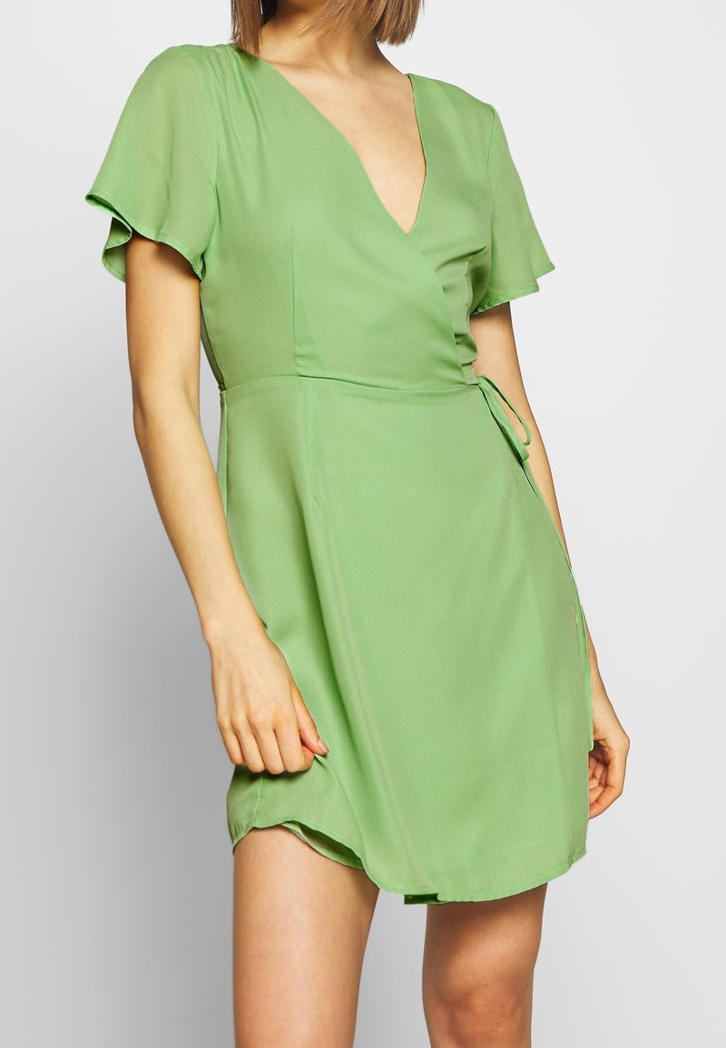 Femme portant une robe portefeuille courte vert clair à manches courtes et col en V, debout devant un fond gris clair uni.