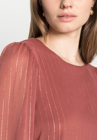 Blusa bordeaux com riscas douradas. Tecido transparente com textura suave. Mangas bufantes franzidas. Primeiro plano do decote e dos brincos usados.