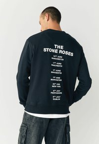 Marineblå sweatshirt med hvid tekst på ryggen, der angiver koncertdatoer og -steder, med rund halsudskæring og ribbede manchetter.