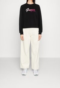 Persona che indossa una felpa nera con il testo rosa "Guess Est. 1981", pantaloni bianchi larghi e scarpe da ginnastica bianche su sfondo semplice.