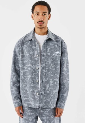 Uomo con tatuaggi sul viso che indossa una giacca grigia con motivo paisley e pantaloni abbinati, in piedi davanti a uno sfondo bianco uniforme.