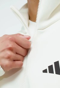 White zip-up jacket with textured fabric, featuring a high collar and black adidas logo on the chest. Zipper is partially closed.