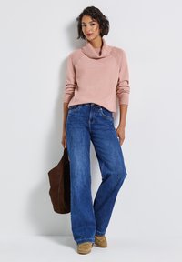Suéter rosa claro con cuello de cisne y hombros texturizados, combinado con jeans de pierna ancha azules y un bolso de ante marrón, sobre un fondo claro.