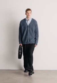 Μπλε υφασμένο κασκόλ με V-λαιμόκοψη, κουμπωτό μπροστά, φορεμένο πάνω από ένα ανοιχτό μπλε ριγέ πουκάμισο. Μαύρο παντελόνι και μαύρη τσάντα στο χέρι.