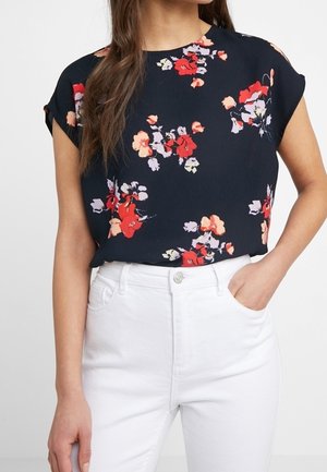 Femme portant un chemisier bleu marine avec des motifs floraux rouges et pêche, associé à un jean blanc taille haute, une main dans la poche.