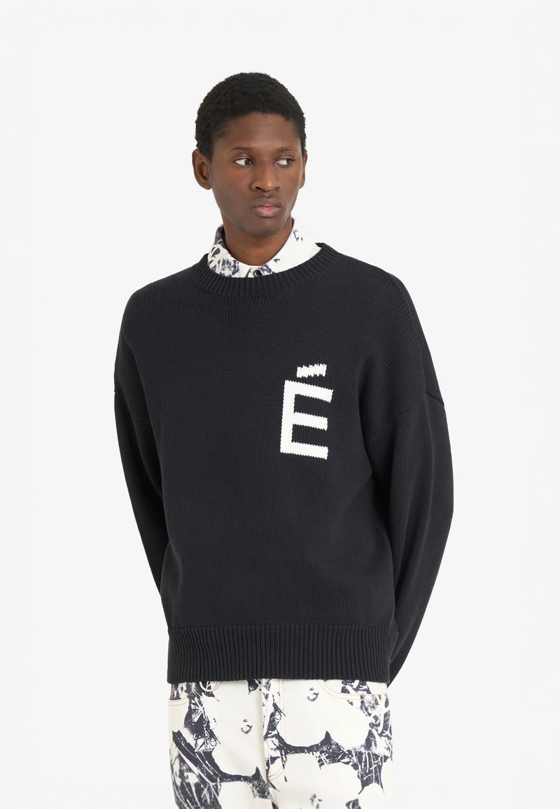 Sweater noir en maille avec le logo blanc "É" sur le devant, encolure ronde et épaules tombantes. Porté par-dessus une chemise à motifs.